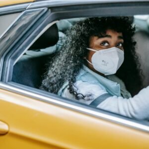Thoughtful African American female with dark curly hair in medical protective mask sitting in taxi and looking away in daytime